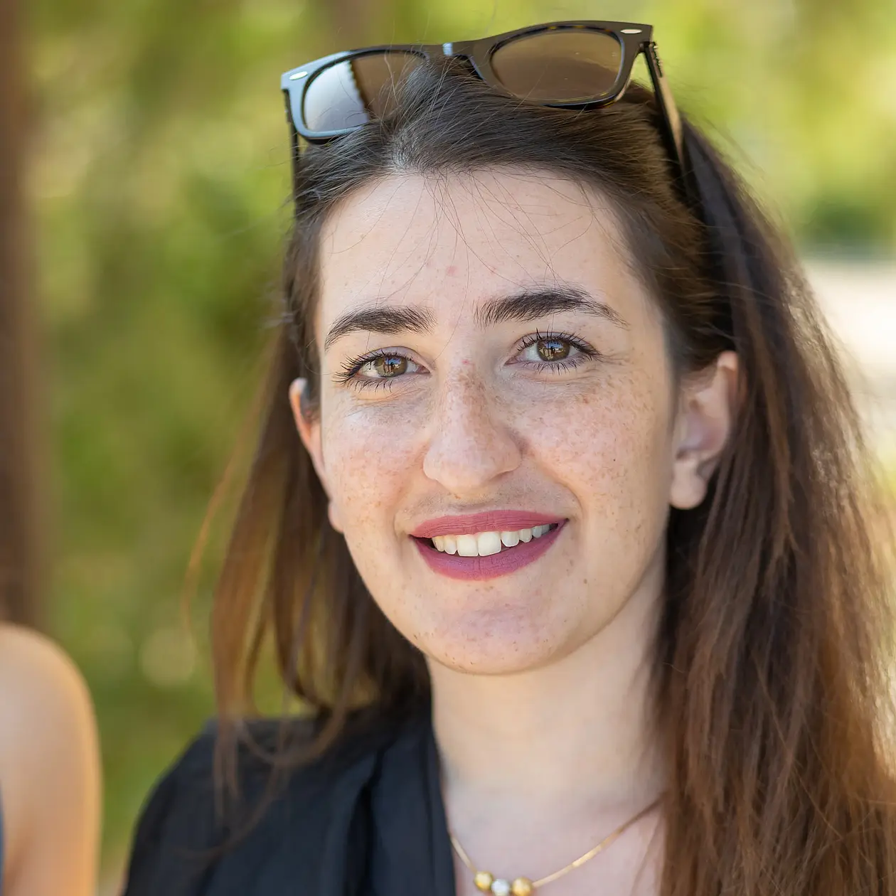 A portrait of Charoula Fotiadou, a white woman with long brown hair an freckles smiling into the camera. She is wearing sunglasses on top of her head and a black blouse as well a golden necklace