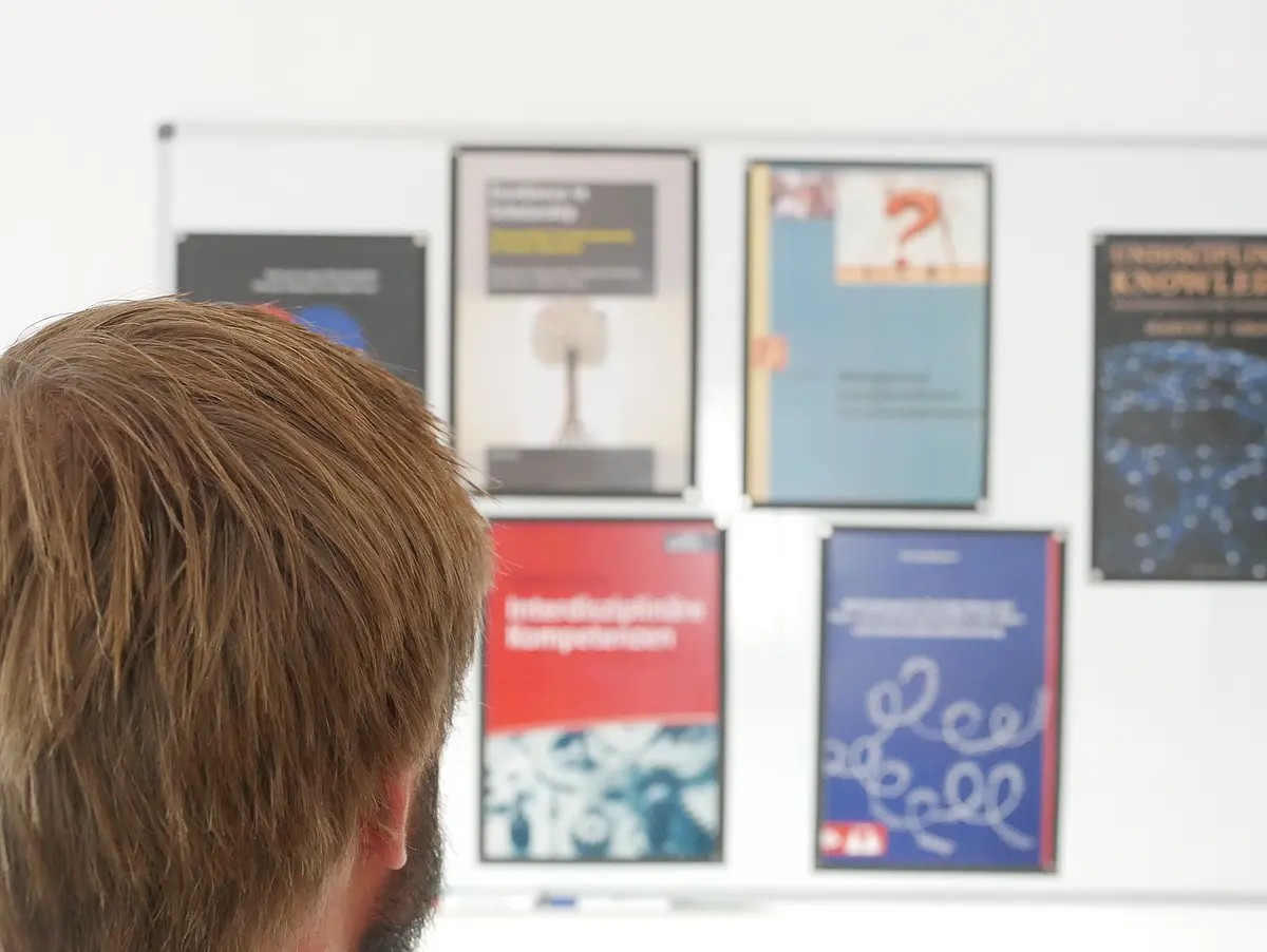 Ein Mensch steht vor einer Wand, an der Titelseiten von Büchern über integrierte Forschung ausgestellt sind. Auf jeder der Seiten ist ein anderes Bild zu sehen, das die Zusammenarbeit in interdisziplinären Teams darstellen sollen. Der Mensch sieht die Bilder an und denkt über sie nach. 