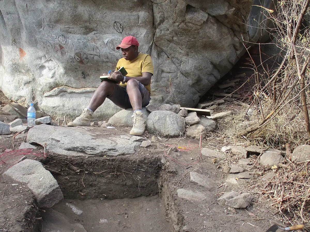 A picture of a fieldwork site, a trench in the foreground with stones and excavation tools like trowels and a bucket in it. In the background, there is a boulder with rock carvings. In front of the boulder, Edward Masongo is sitting holding a folder and writing. He is a black man wearing a red baseball cap, a mustard yellow short sleeve shirt and short cargo pants as well as beige desert working boots.  