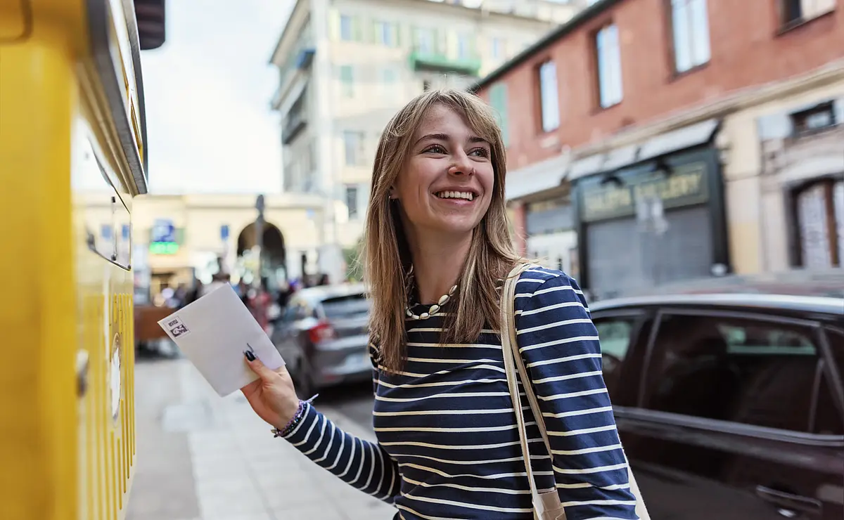 Frau auf der Straße mit einem Brief in der Hand, den sie in einen Briefkasten wirft.