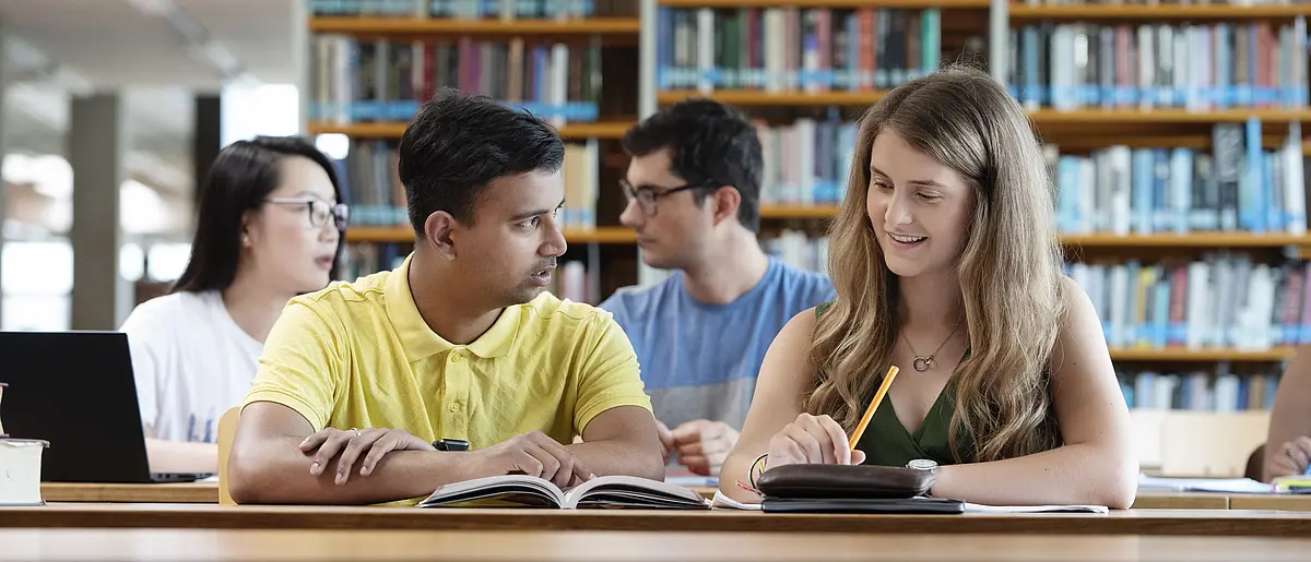 Vier Studierende (2 Frauen und 2 Männer) sitzen in der Bibliothek und besprechen die zu lösenden Aufgaben. 