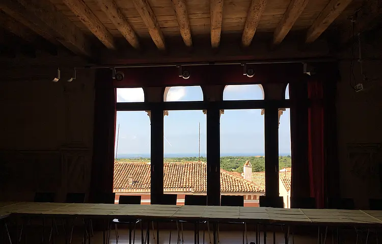 Blick aus vier hohen Fenstern mit Sprossen unterteilt, davor eine Tischreihe mit Stühlen. Das Bild wurde bei einer Konferenz in Bale aufgenommen.