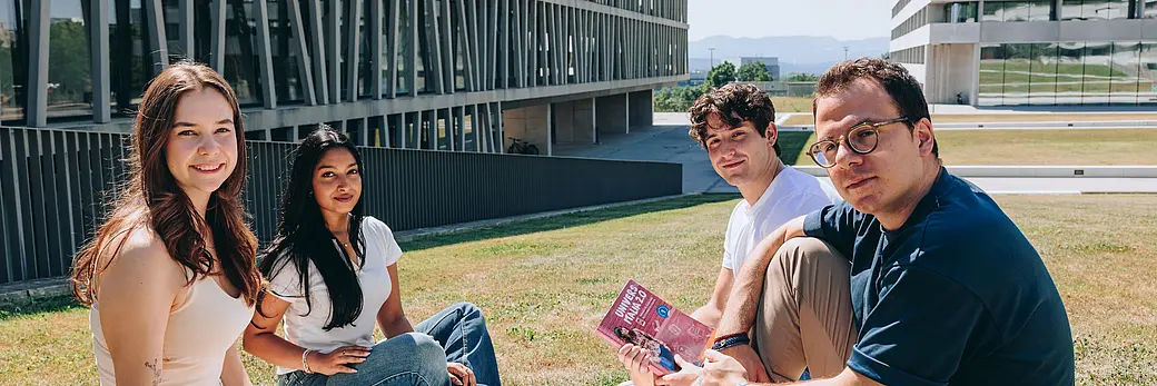 Four students are sitting on a lawn in front of a lecture hall building