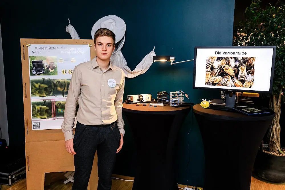 Der Sieger Sebastian Steppuhn steht links im Bild vor einer Infotafel, auf der ein Poster angeheftet ist. Rechts im Bild steht ein Computerbildschirm, auf dem Bienen gezeigt werden mit der Überschrift "Die Varroamilbe". Im Hintergrund ist ein Imkeranzug an der Wand aufgehängt. 