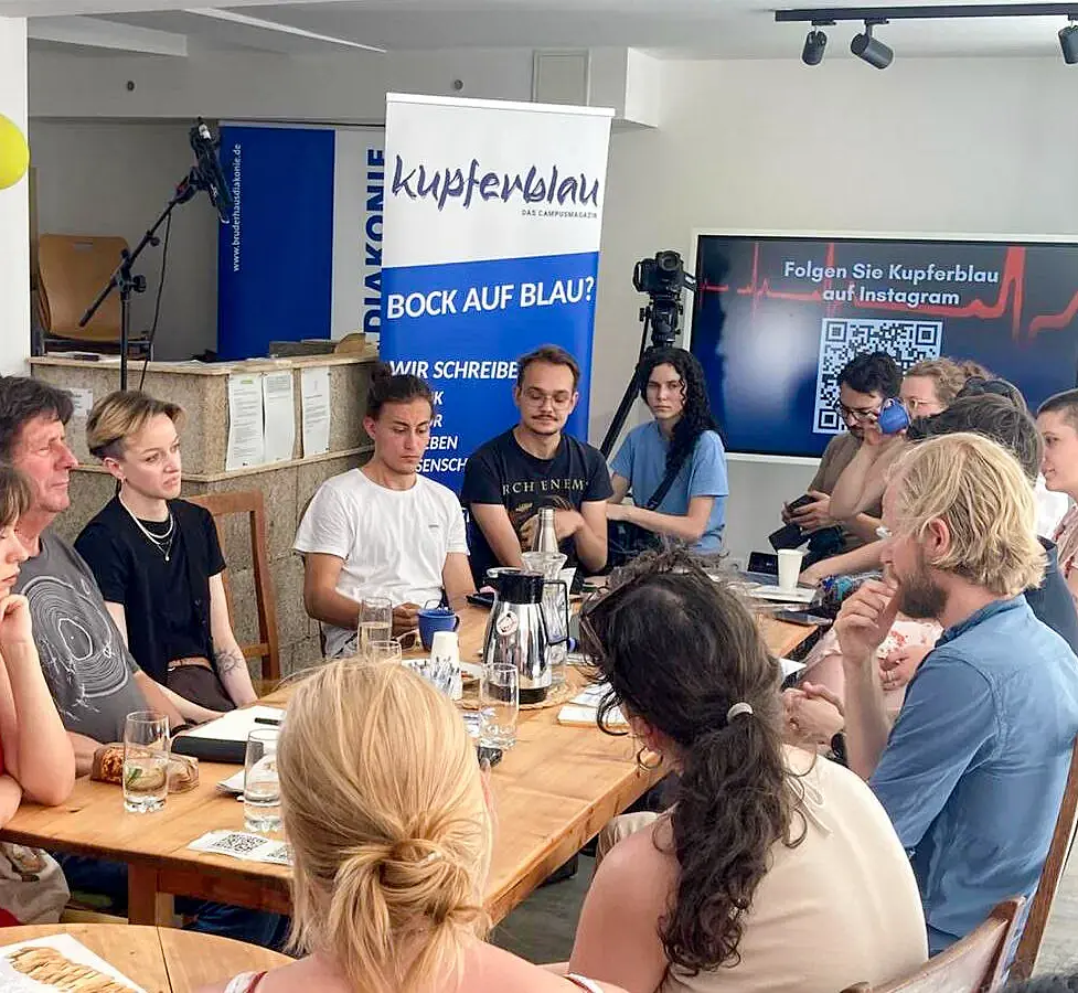 Ein Gruppe von ungefähr 10 jungen und älteren Personen sitzt um einen Tisch und diskutiert miteinander. Im Hintergrund steht ein Aufsteller des Campusmagazins kupferblau.