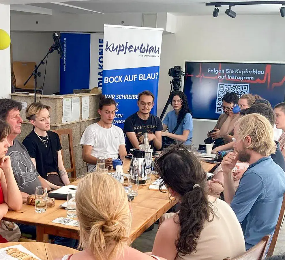 A group of about 10 young and older people are sitting around a table and chatting. In the background, there's a display stand for the campus magazine kupferblau.