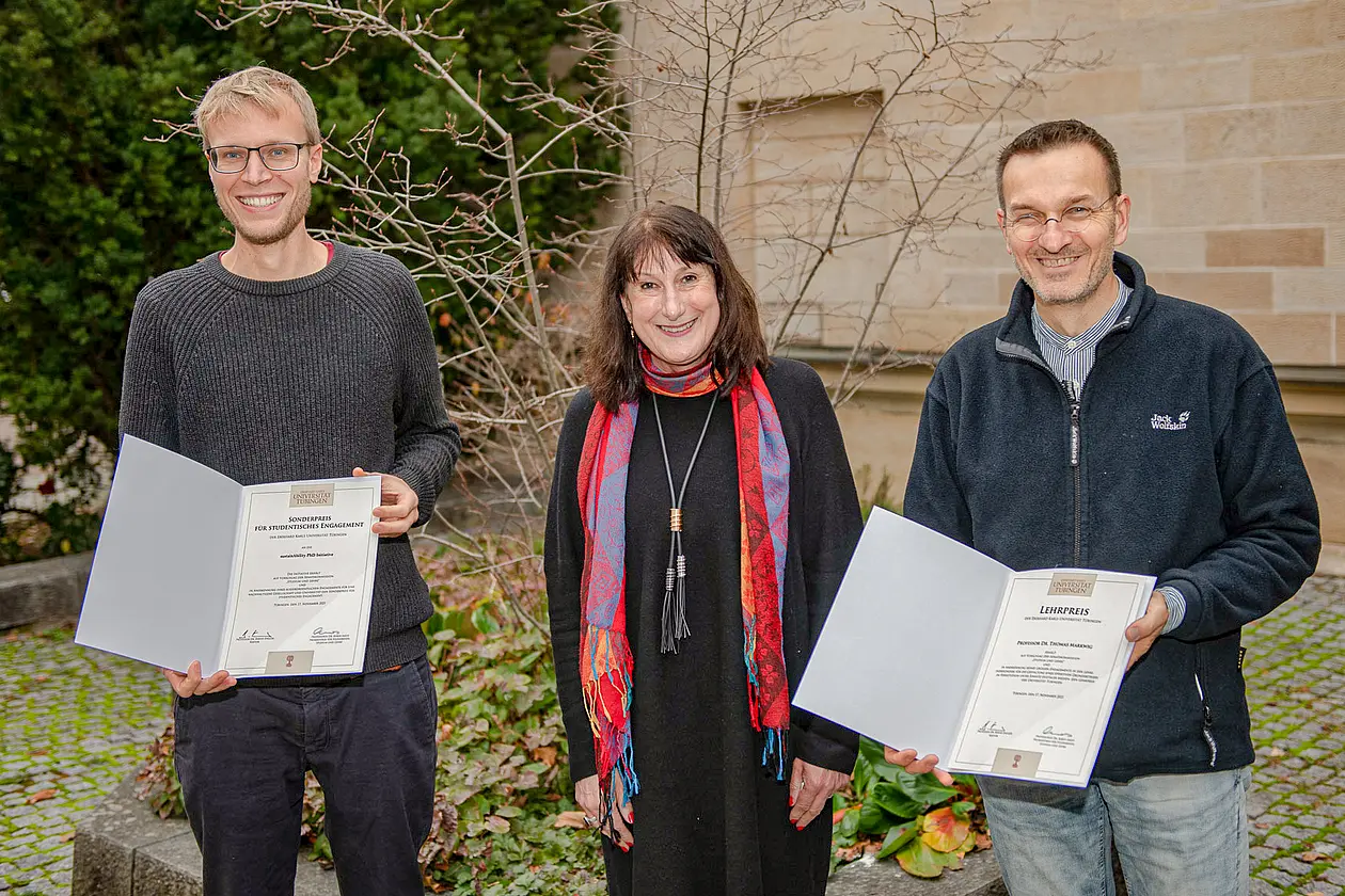 Cornelius Schröder und Thomas Markwig stehen rechts und links von Karin Amos, beide präsentieren eine Urkunde