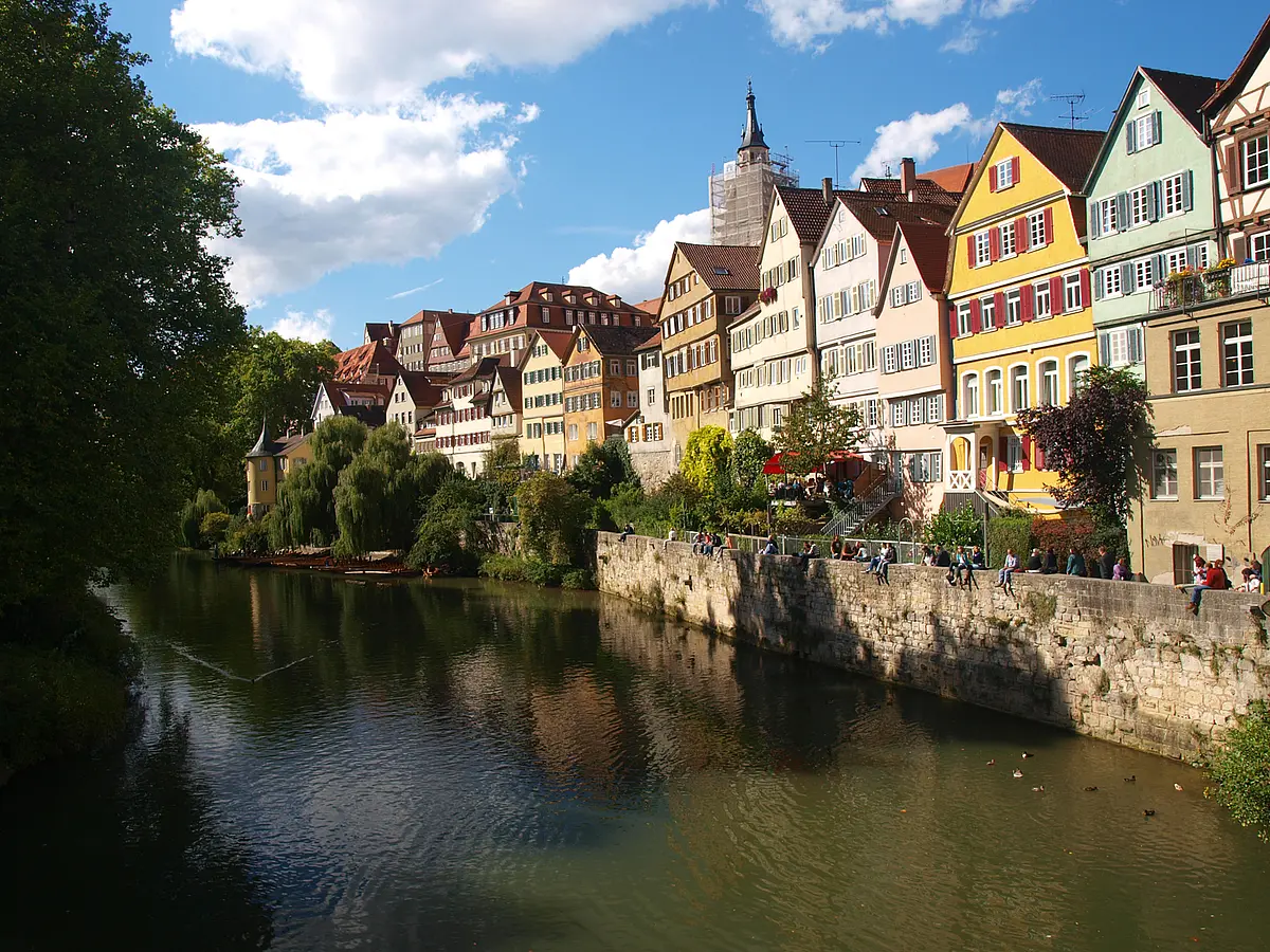 This picture shows the famous Neckar Front, a series of half-timbered houses overlooking the Neckar. Along the Neckarfront there is a wall with people sitting on it. On the left side of the picture one can see the treetops of the poplars on the Neckar island.