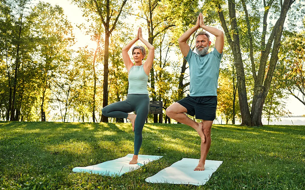 Eine Frau und ein Mann, die Yoga praktizieren auf einer Matte in einem Park. 