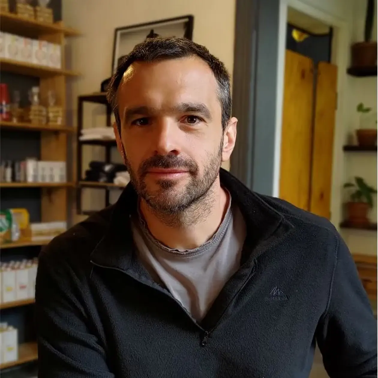 Portrait Photo of Patrick Schmidt, a white man with very short black hair and beard stubbles, wearing a dark blue sweater and a grey shirt, sitting in a room with large wooden displays
