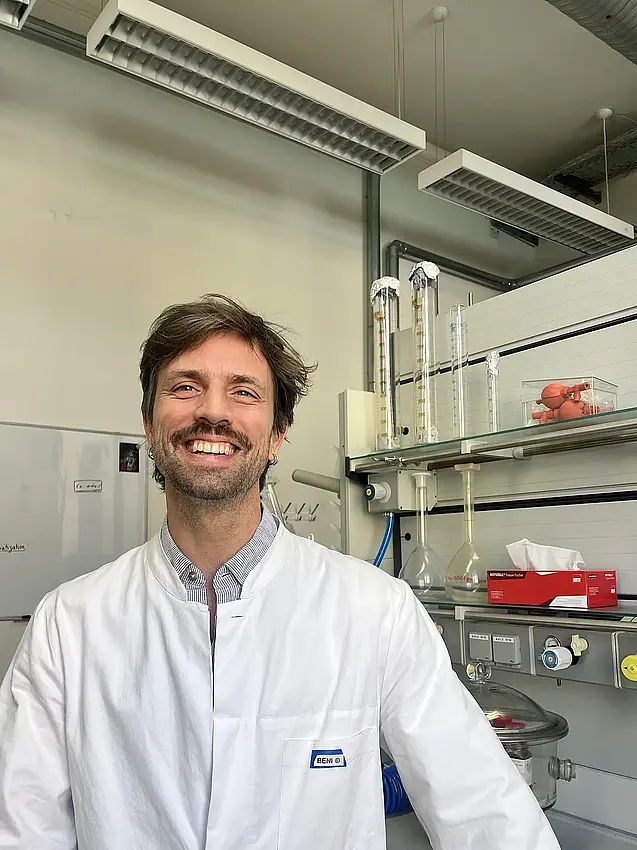 A photo of Benjami Laubi, a white man with a shaggy brown haircut, a schnauzer and beard stubbles, wearing a blue and white caro buttondown under a white lab coat. He smiles widely at the camera while standing in a lab environment with glasses and machinery behind him. 