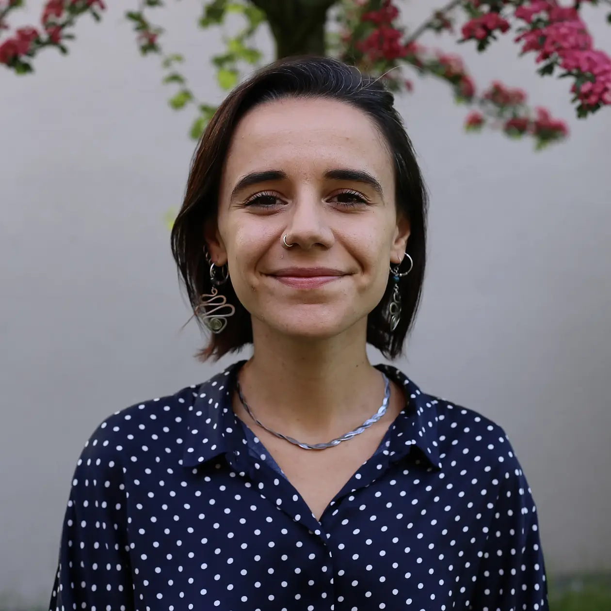 A photo showing Cecilia Padilla-Iglesias from the front. She is a white woman with a mediterranean type with dark brown hair in a short bob, smiling widely at the camera. She wears a dark blue blouse with white polka-dots and silver necklace, as well as large, snakelike silver earrings. She stands in front of a small tree with bright pink blossoms.