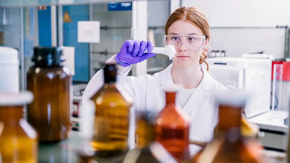 Eine Schülerin im weißen Laborkittel prüft einen Wattebausch mit chemischer Flüssigkeit vor dem Regal eines Laborraums. Schülerlabornetzwerk MINT me!