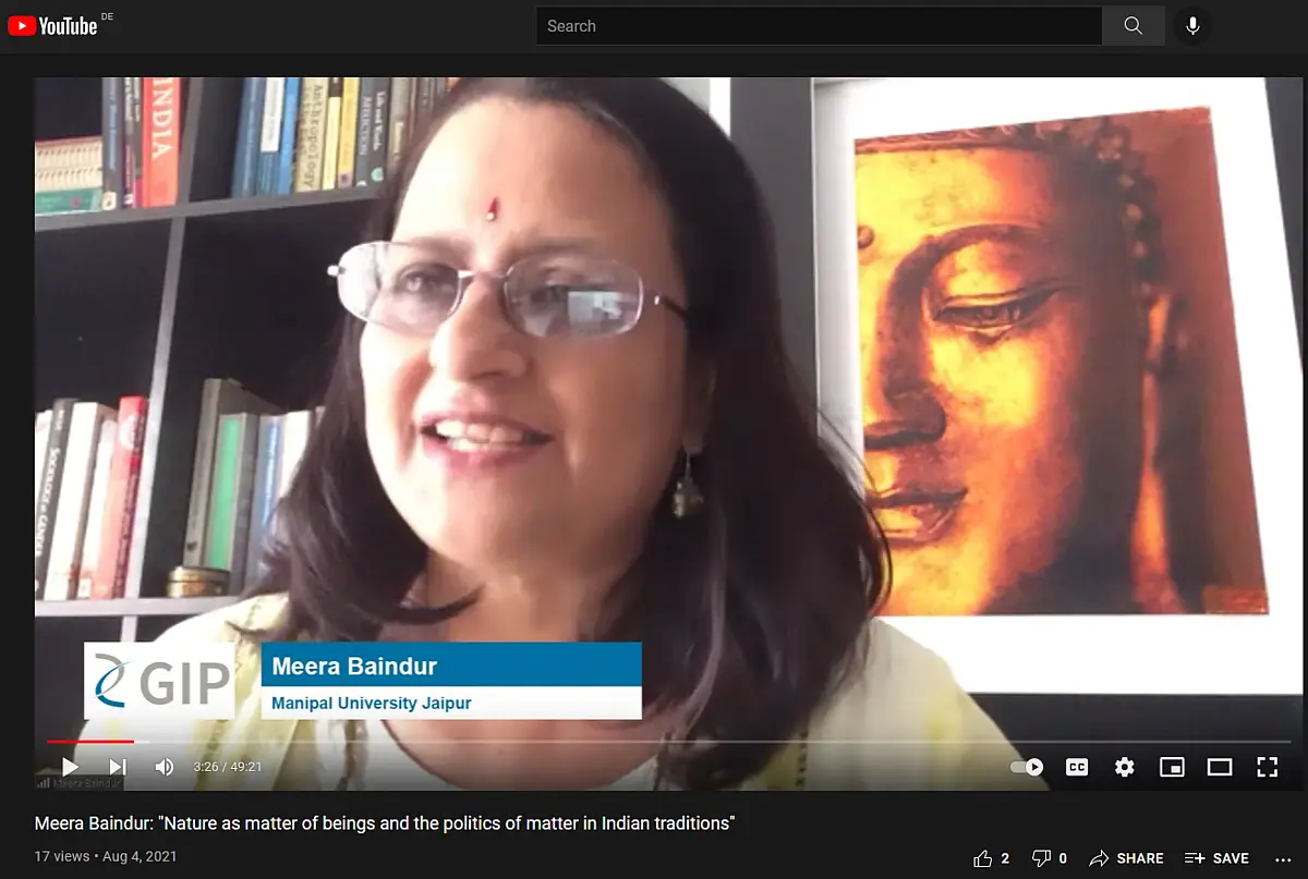 A woman with dark hair speaks into a webcam, in the background hangs a Buddha picture