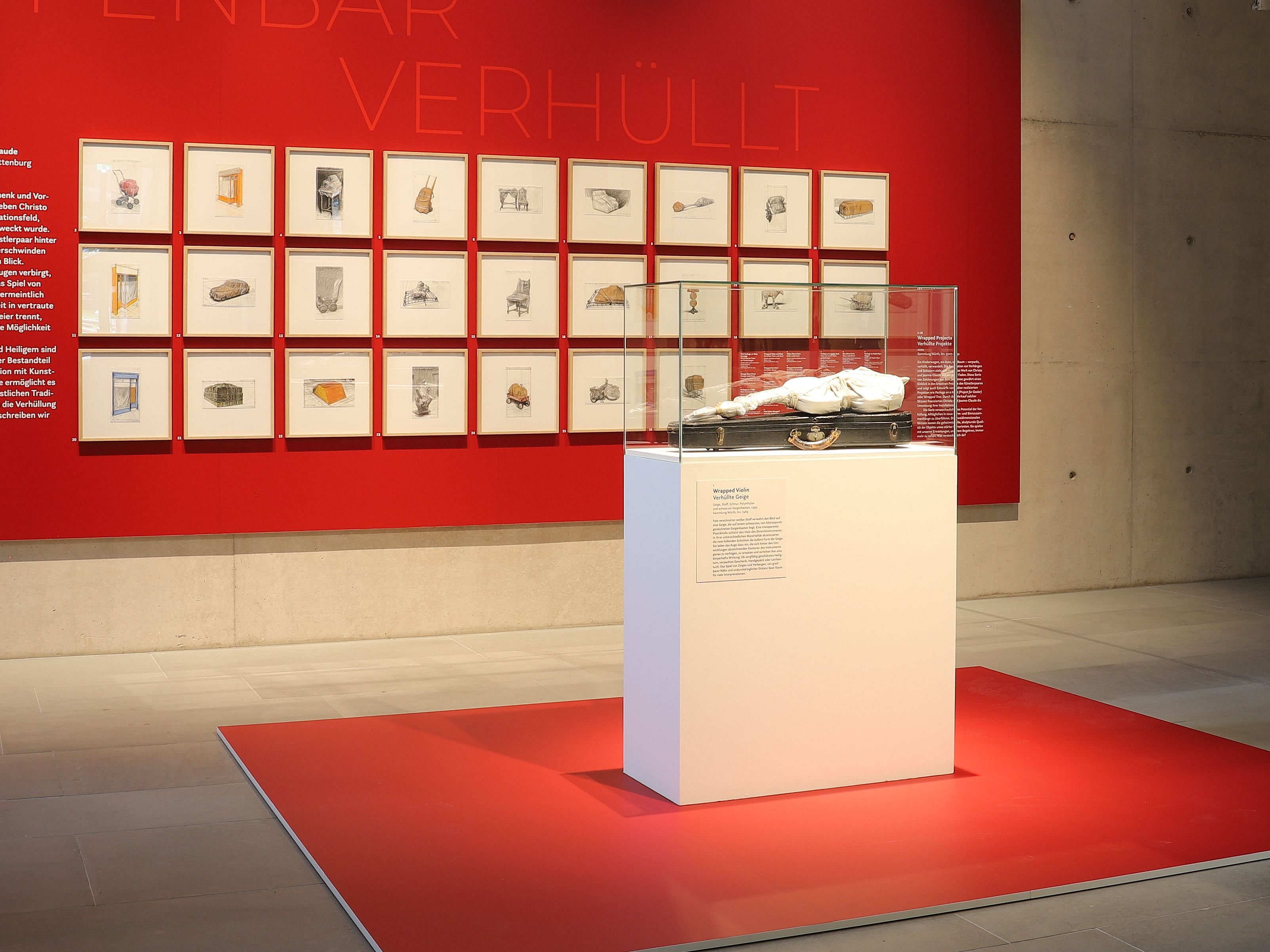 Foto der "Verhüllten Geige" Christos im Diözesanmuseum Rottenburg vor dem Hintergrund von 24 Zeichnungen Christos, eingerahmt vor rotem Hintergrund
