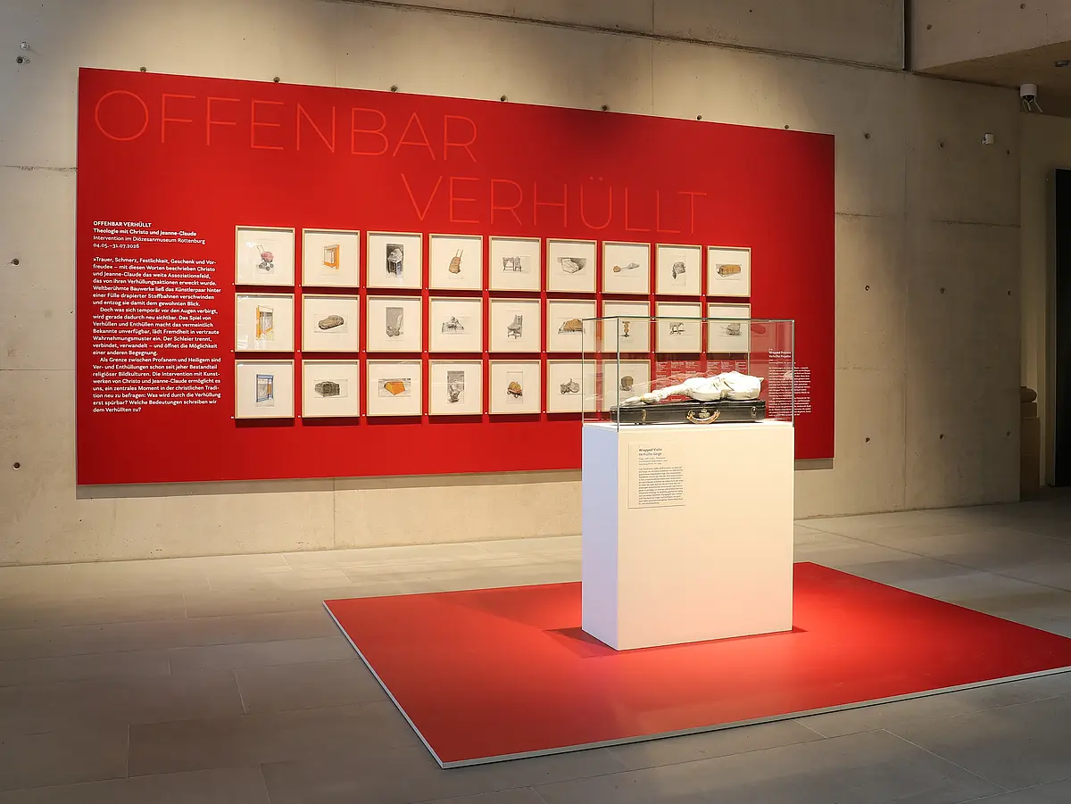 Foto der "Verhüllten Geige" Christos im Diözesanmuseum Rottenburg vor dem Hintergrund von 24 Zeichnungen Christos, eingerahmt vor rotem Hintergrund