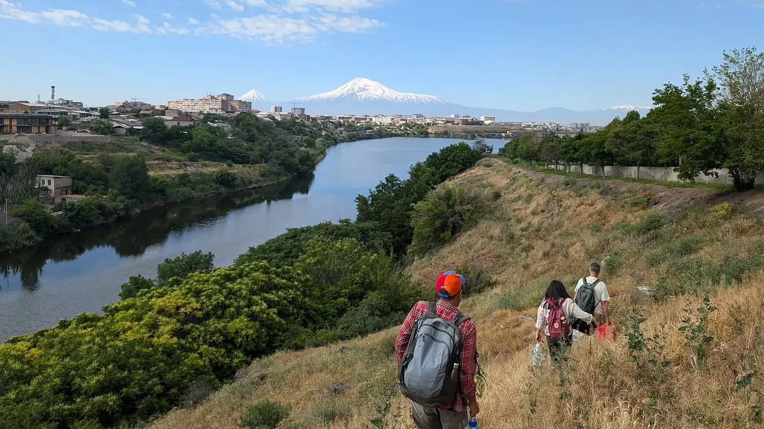 Photo of a river, with a city to the left of it and three people with backpacks out in the countryside to the right of it