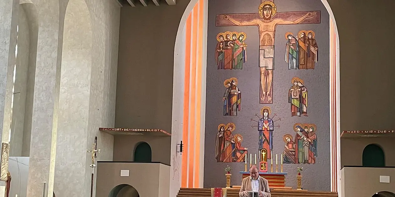 Dieses Bild wurde aus einer Kirchenbank aufgenommen. Mehrere Menschen sitzen offenbar in den Bänken. Am Ambo steht ein älterer Herr mit Halbglatze und weißen Haaren und einer Hornbrille. Der Mann trägt etwas vor.