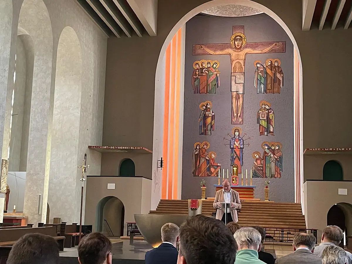 Dieses Bild wurde aus einer Kirchenbank aufgenommen. Mehrere Menschen sitzen offenbar in den Bänken. Am Ambo steht ein älterer Herr mit Halbglatze und weißen Haaren und einer Hornbrille. Der Mann trägt etwas vor.