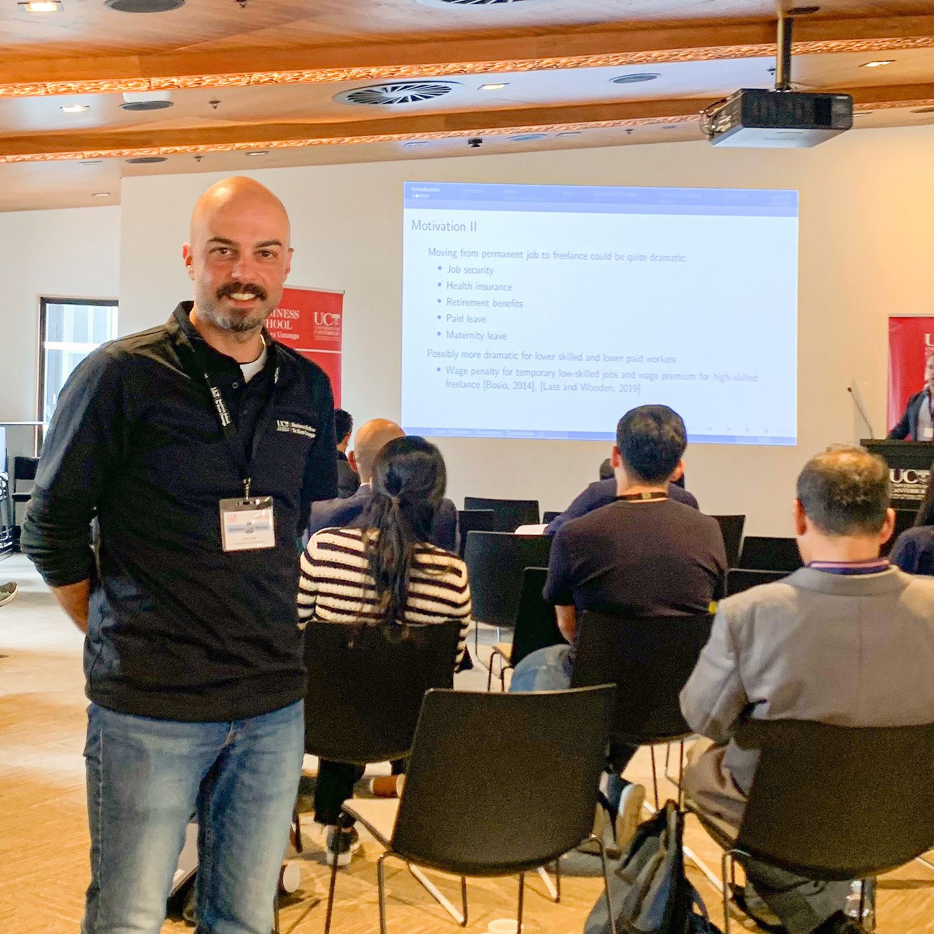 Dr. Onur Kıska standing on the left of the picture at the 17th Australasian Trade Workshop. Behind him, a presentation slide is displayed on a large screen, and several chairs with conference attendees are visible in the background.