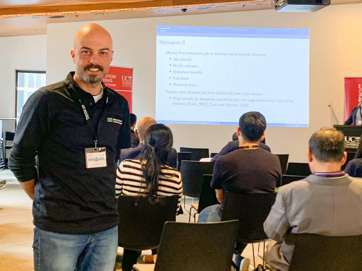 Dr. Onur Kıska standing on the left of the picture at the 17th Australasian Trade Workshop. Behind him, a presentation slide is displayed on a large screen, and several chairs with conference attendees are visible in the background.