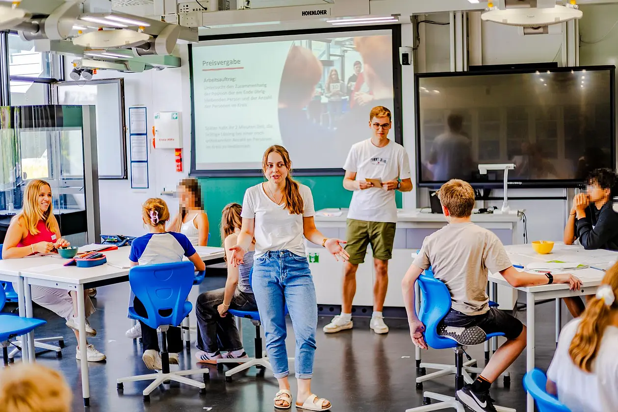 Zwei Studierende als Lehrende im Klassenzimmer, Schüler in Sitzgruppen