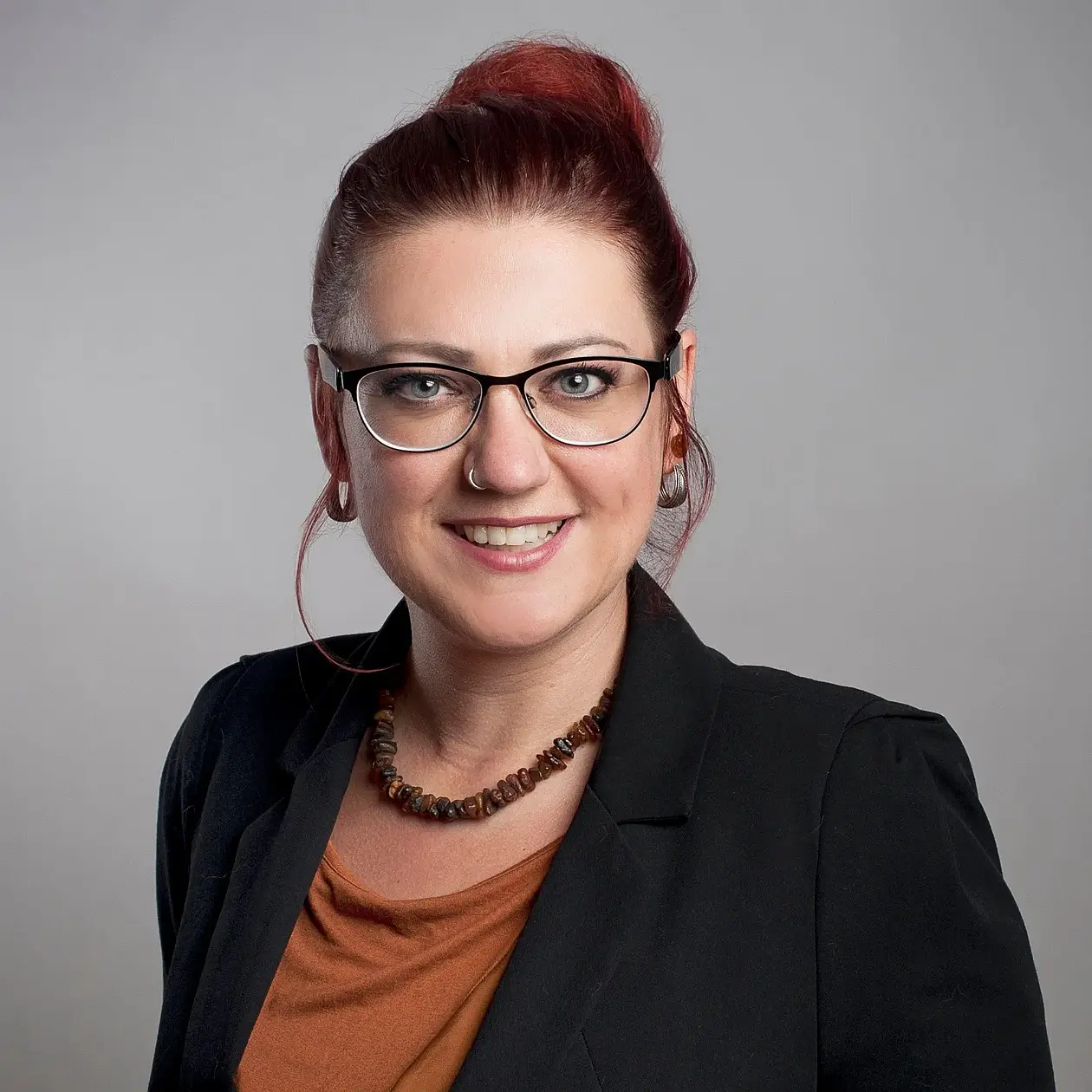 A photo of Nicola Scheyhing, a middle aged white woman with red hair in an updo and glasses, wearing a black blazer and an amber brown shirt as well as an amber necklace. She is facing the camera in front of a middle grey screen. 