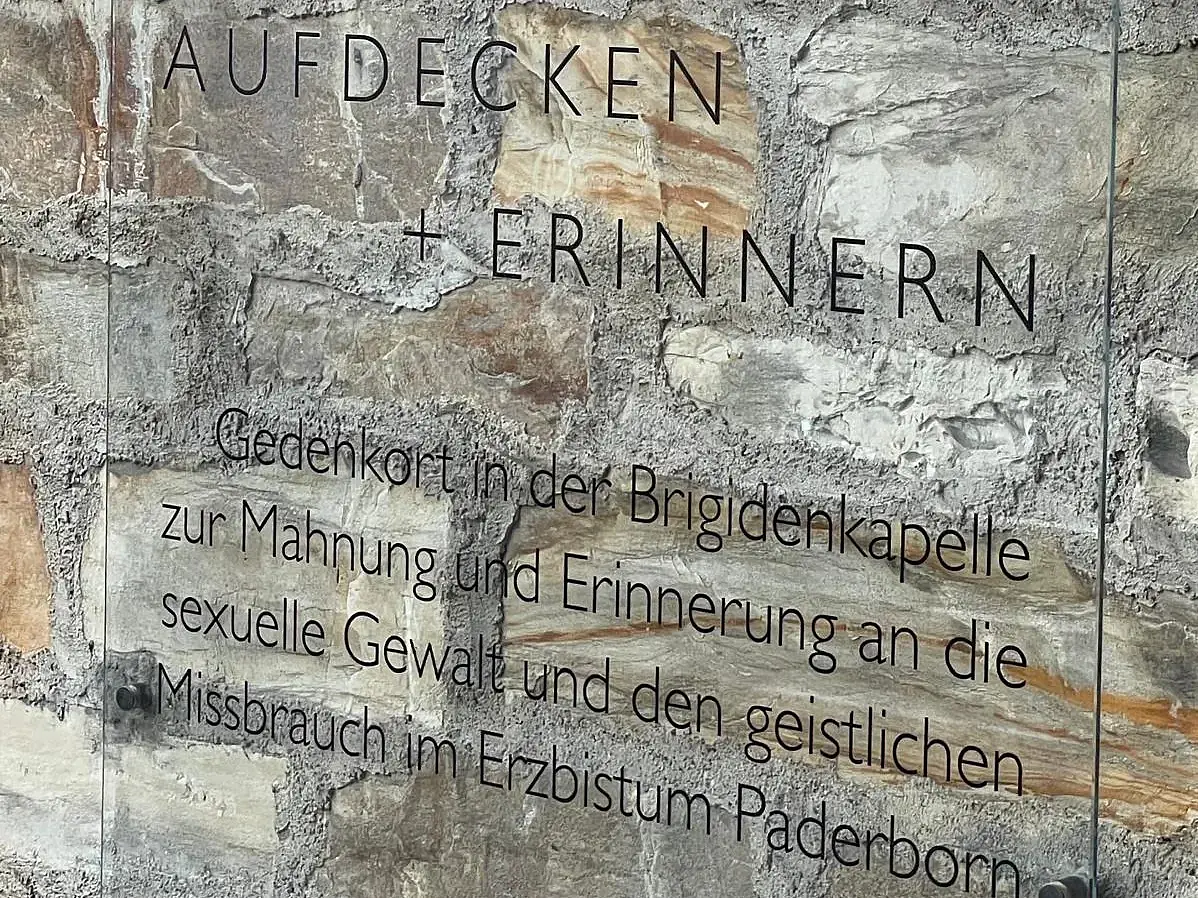 Zu sehen ist eine Glastafel, die an eine steinerne Wand angebracht ist. Auf ihr steht: "Aufdecken und Erinnern. Gedenkort in der Brigidenkapelle zur Mahnung und Erinnerung an die sexuelle Gewalt und den geistlichen Missbrauch im Erzbistum Paderborn"