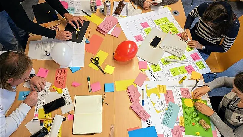 Bunte Zettel, Post-its und Bastelmaterial liegen auf einem Tisch, um den herum Menschen sitzen. Von der Idee zur Wirkung