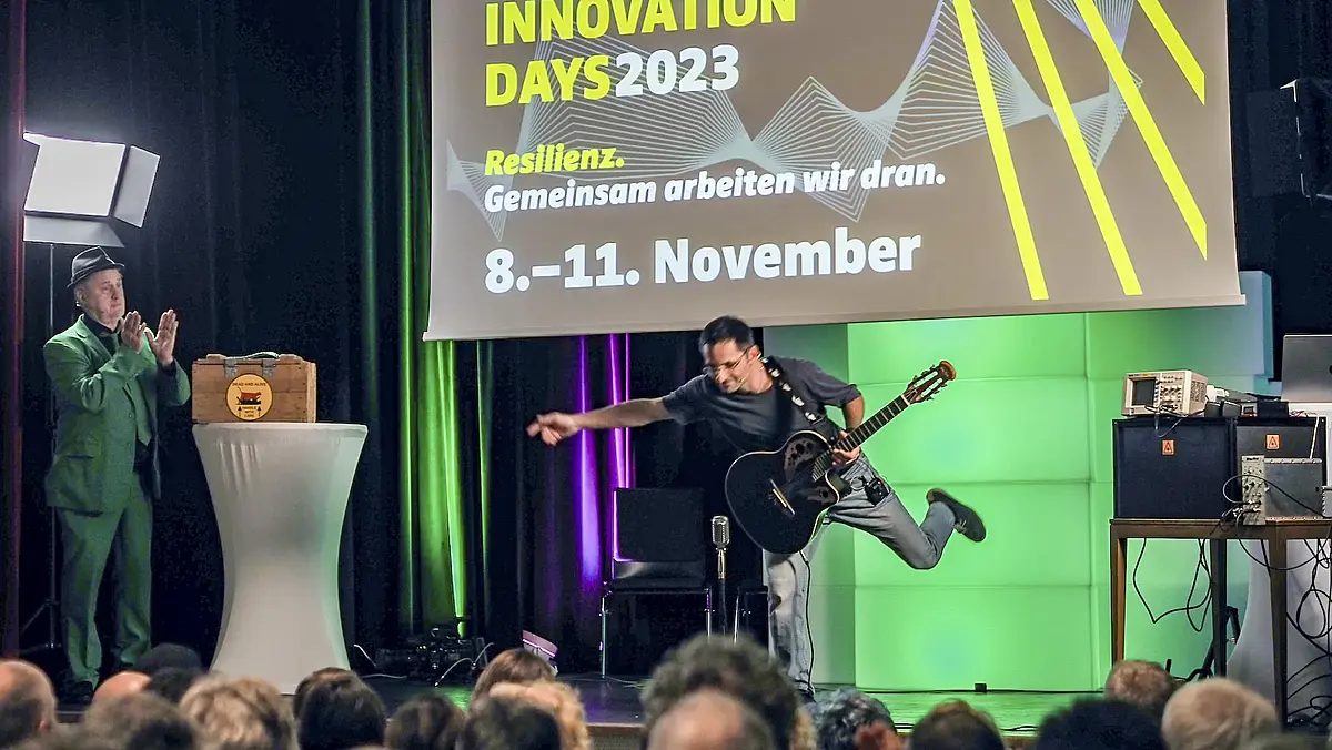 Auf einer Bühne der Science & Innovation Days sieht man einen Mann mit einer Gitarre, der sich verbeugt und einen Mann mit grünen Anzug und Hut, der klatscht. Hörsaal 7