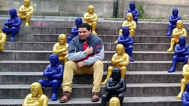 Junger Mann Mitte 30 mit dunklen Haaren, der auf einer Treppe neben gelben und blauen Skulpturen sitzt