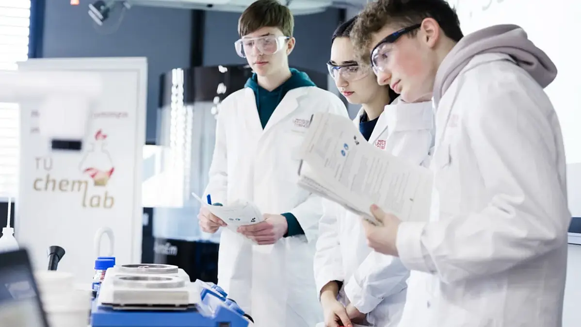 Zwei Schüler und eine Schülerin der Oberstufe in weißen Laborkitteln und mit Schutzbrillen prüfen eine chemische Versuchsanordnung im TüChemLab.