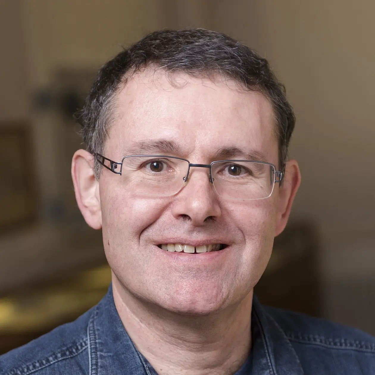 A portrait photo of Hervé Bucherens, a middle aged white man with short curly grey hair and rectangular glasses, smiling at the camera, wearing a demin button down shirt. 