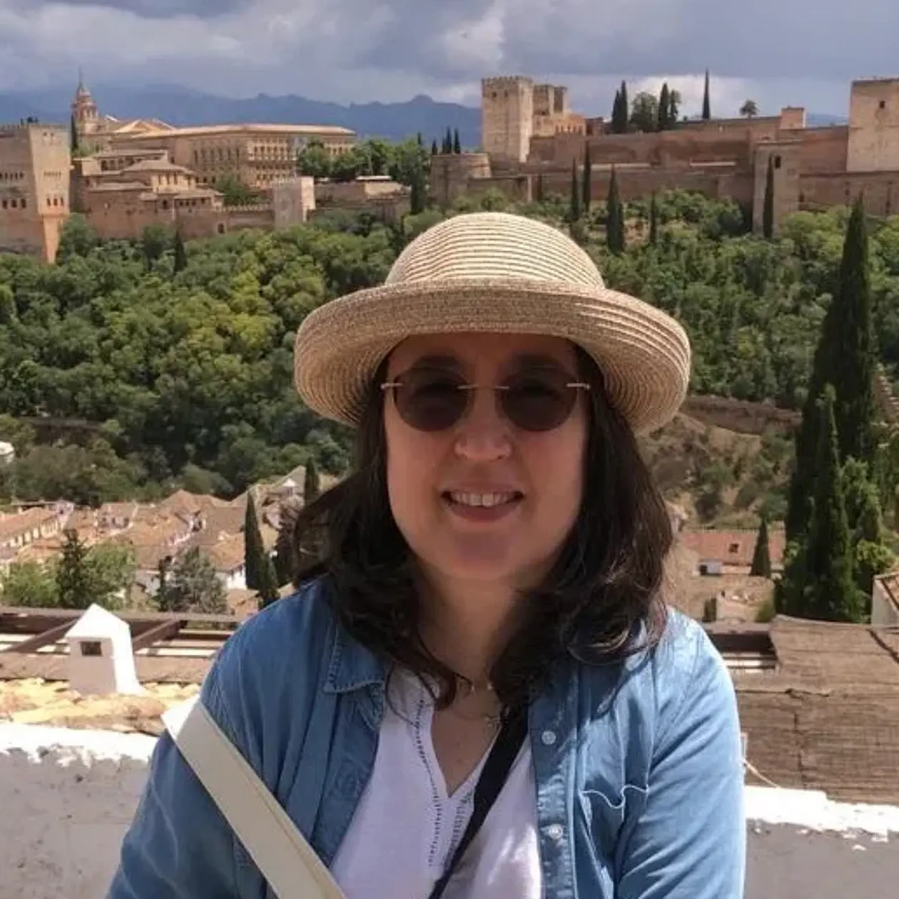 A photo of Dorothée Drucker, a white woman with medium long dark brown hair wearing sunglasses, a straw hat, a light blue cardigan and a white shirt standing in front of the panorama of a medieval mediterranean city. 