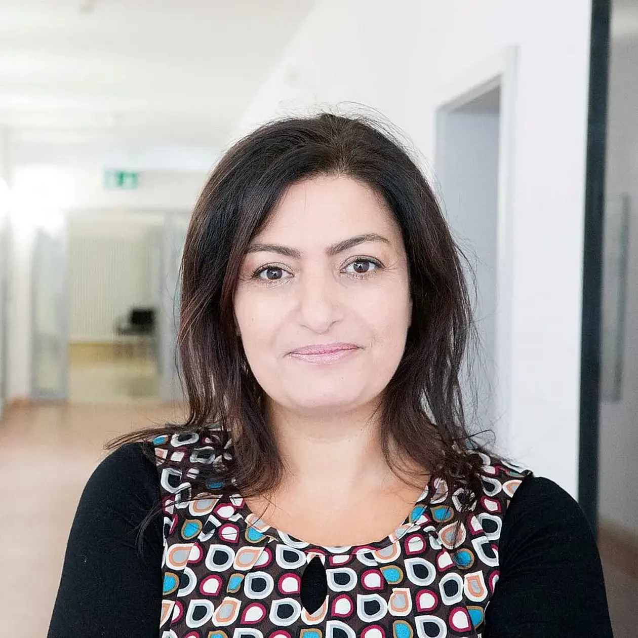 A portrait photo of Sireen el Zaatari, a woman with levantine heritage with long dark brown hair ending at her shoulders, wearing a black Sweatshirt with a geometric pattern on the torso in blue, green, white, brown, resembling stylized leaves. She is standing in a light hallway, facing the camera and smiling. 