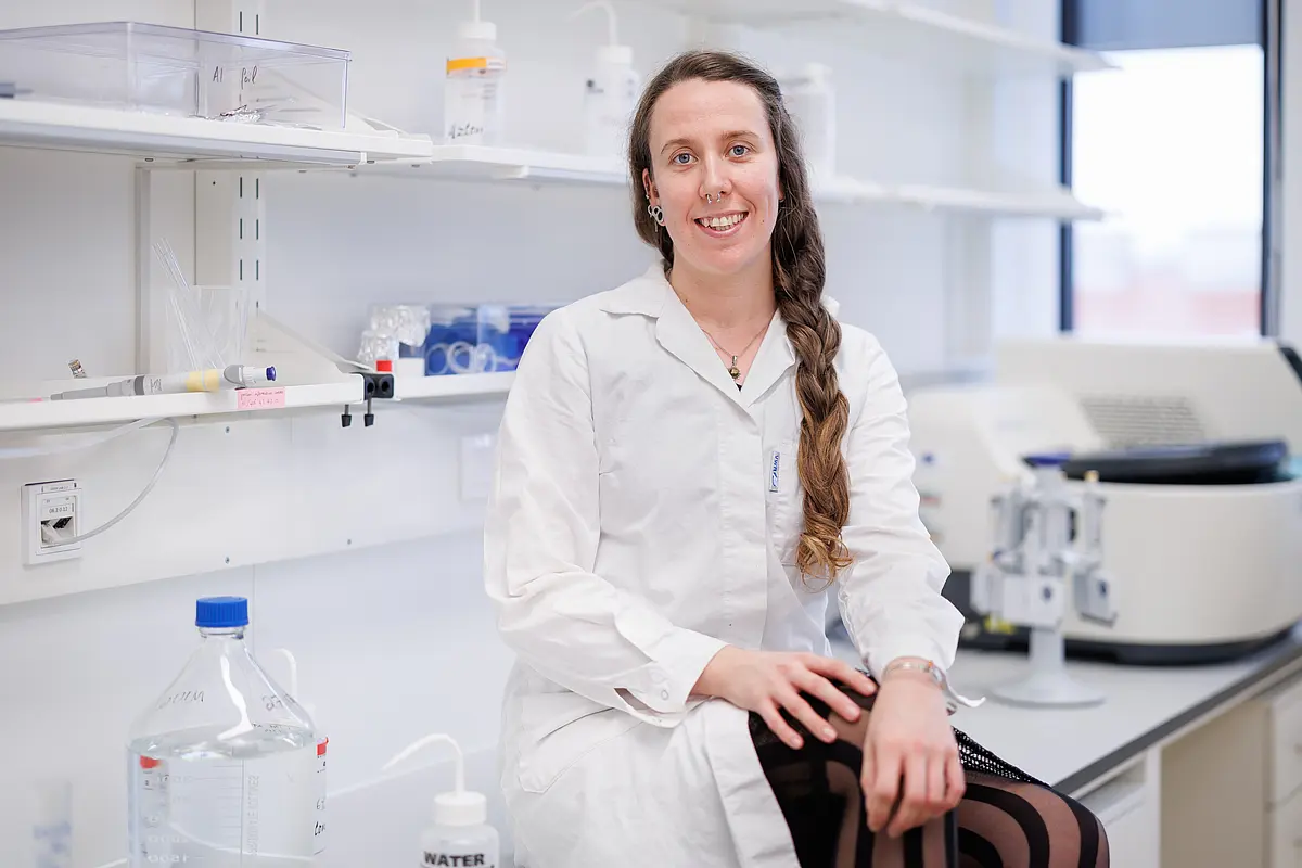 Dr. Emily Pigott, a white woman with long brown hair wearing a white lab coat, is sitting on the table of a lab environment with white cupoards, glass container and machines. She has several piercings in her ear and in her nose septum. 