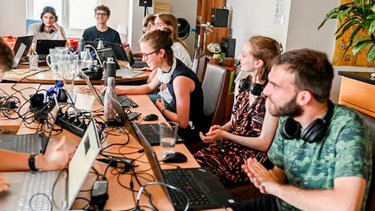 Fröhliche junge Menschen sitzen im Hauptraum des Makerspace vor aufgeklappten Laptops und programmieren. KI-Makerspace