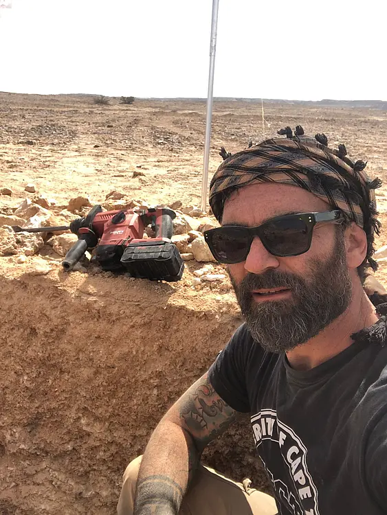 A field photo of Yamandu Hilbert in a trench in an arid landscape. In the background, excavation tools and a measurement pole is visible. Dr. Hilbert is a white man with a mediterranean complexion, a black beard with greys in it, wearing a black shirt with a white graphic print, a black and white headscarf, and black sun glasses.