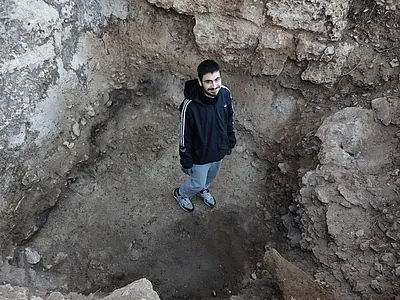 Iasonas Nestoridis, a white man with short black hair and a short black beard with a prominent moustache wearing a black training jacket with a hood and blue jeans, stands in a deep excavation pit. The photo is taken from higher above. 