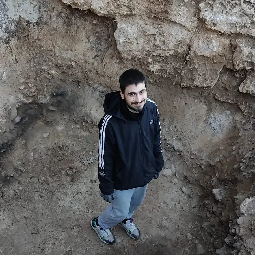 Iasonas Nestoridis, a white man with short black hair and a short black beard with a prominent moustache wearing a black training jacket with a hood and blue jeans, stands in a deep excavation pit. The photo is taken from higher above. 