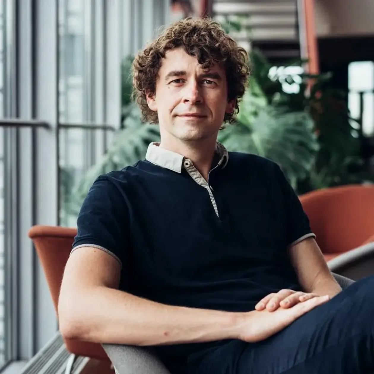 Photo of Johannes Krause, a white man with brown curly hair that is a bit longer on top and combed to one side. He ist sitting in a red lounge chair in front of a window panel with plants in the background, wearing a short sleeve dark blue shirt with a white collar and dark jeans. 