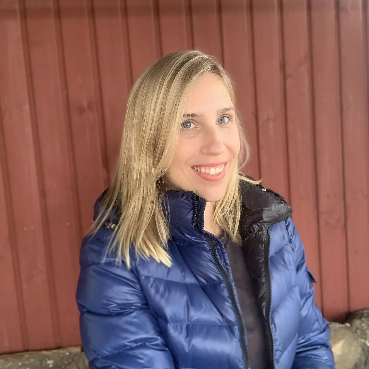 A photo of Melanie Faiss, a white woman with medium long blond hair, wearing a blue puffer jacket, standing in front of a reddish brown wall of wooden panels. 