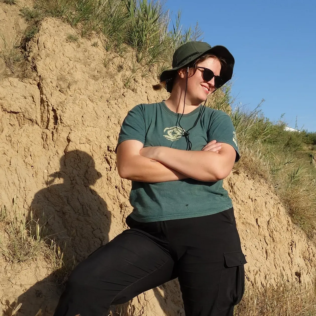 A photo of Tegan Foister, a white woman standing with crossed arms in front of a mud profile. She is wearing black cargo trousers, a teal shirt, dark sunglasses and a wide brim olive hat. 