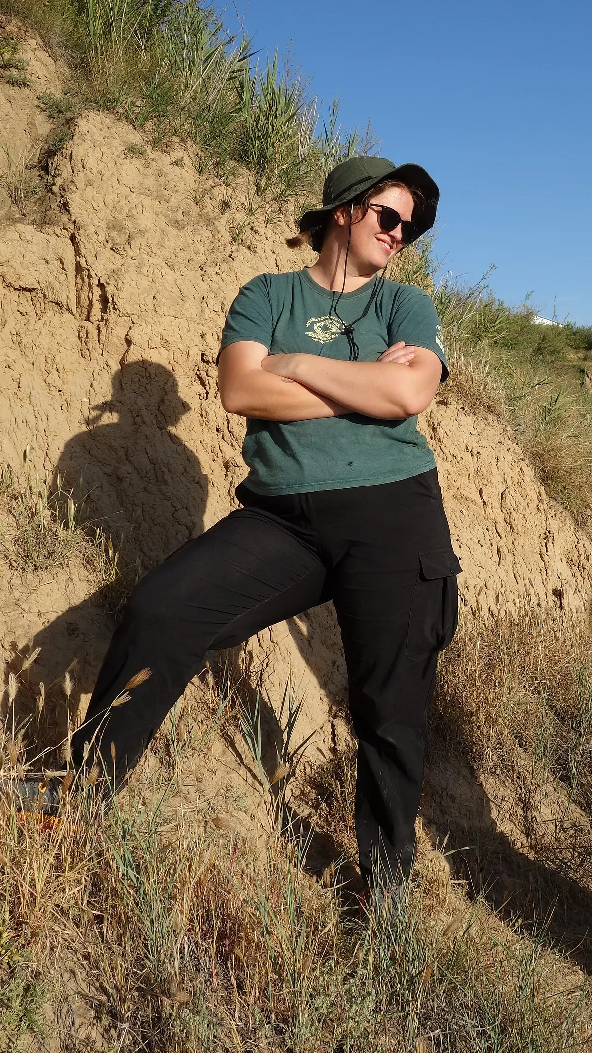 A photo of Tegan Foister, a white woman standing with crossed arms in front of a mud profile in a fieldwork situation. She is looking right, wearing a wide brimmed olive hat, dark sunglasses, a teal shirt and black cargo pants. 