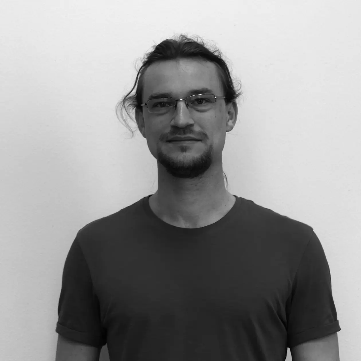 A black and white photo of David Kirschenheuter, a white man with long dark hair in a ponytail and a short beard, wearing a dark T-Shirt and rectangular glasses. 