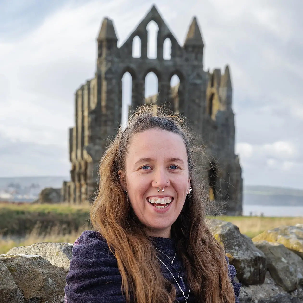 The photo of Emily Pigott shows a white woman with long brown hair in a half up ponytail smiling into the camera in front of the ruin pf a medieval church in a grassy landscape. She is wearing a dark blue running jacket. 