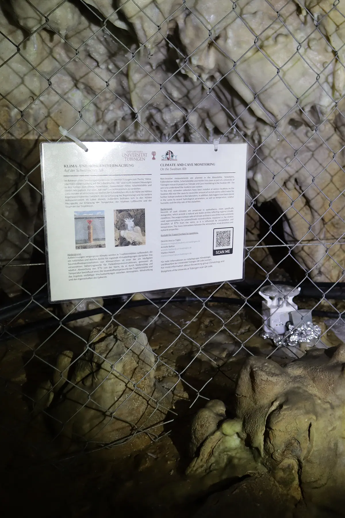 An einem Maschendrahtzaun in einer Höhle ist eine Informationstafel befestigt. Hinter dem Zaun sind Logger zu sehen unter Tropfsteinen.
