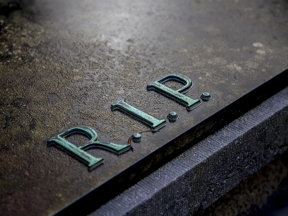 Auf dem Bild ist der Ausdruck "R.I.P." als Messing-Verzierung auf einem dunklen Stein zu sehen.