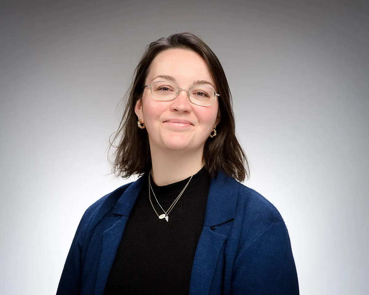Portraitfoto von Frau Dr. Laura Moncion vor einem neutralen Hintergrund, lächelnd frontal in die Kamera schauend, formal in blau und schwarz gekleidet, schulterlange dunkle Haare und Brille