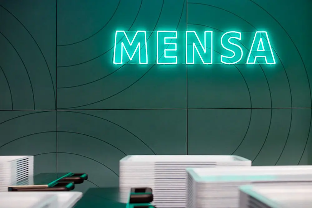 Eine grün leuchtende Mensa-Wandreklame. Davor Mensatabletts, bereit zum Befüllen.